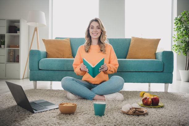 漂亮年轻女孩的全身照片坐在地板上看书学习零食穿橙色的衣服现代室内平坦的室内图片下载