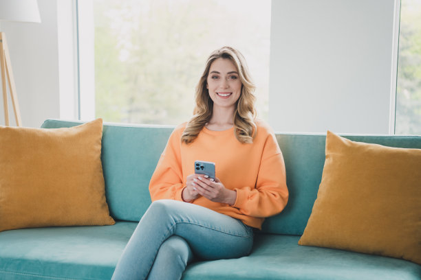 漂亮开朗女孩的照片肖像坐在沙发上拿着小玩意穿着橙色的衣服在宽敞的室内房间里消磨时光图片下载