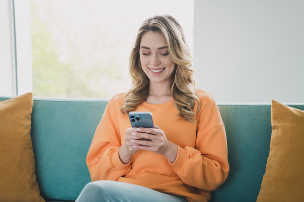 漂亮的年轻女孩的照片肖像坐在沙发上拿着装置穿着橙色的衣服现代室内平坦的室内图片下载