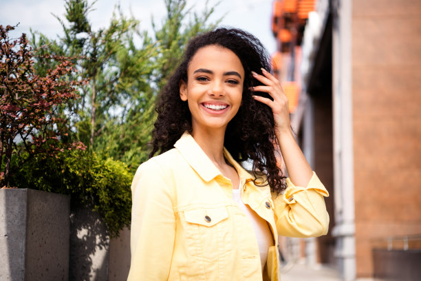 年轻快乐的少女抚摸卷发的照片，学生穿着黄色夹克，在最喜欢的家乡城市户外消磨时光图片下载