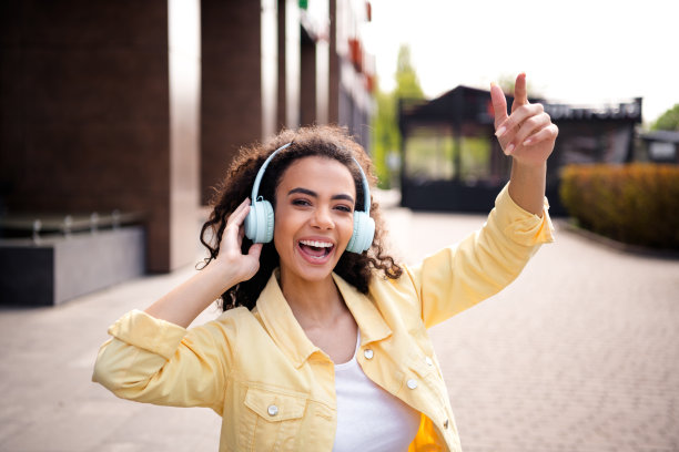 年轻的积极女孩的照片，波浪发型，学生穿着黄色时尚夹克，戴着耳机，在阳光下度过周末图片下载