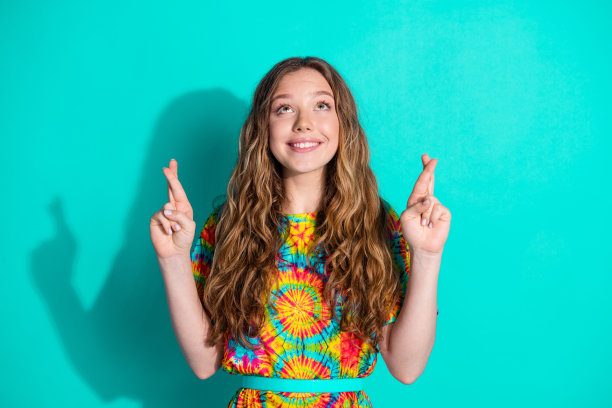 漂亮的十几岁女孩的照片，交叉手指许愿，仰望空旷的空间，穿着时髦的彩色服装，孤立在海蓝宝石的背景上图片下载