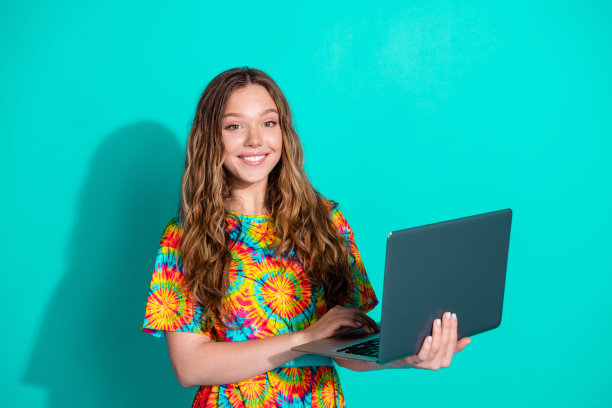 漂亮的十几岁的女孩的照片肖像拿上网本穿时髦的彩色服装孤立的海蓝宝石色背景图片下载