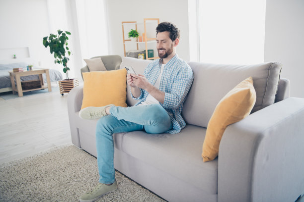 英俊的大胡子的成年男子独自呆在家里的客厅与巨大的沙发阁楼设计和浏览新闻电话的照片图片下载