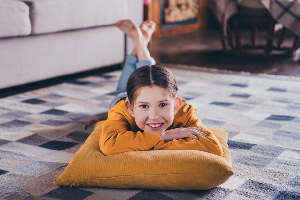 迷人的小女孩躺在地板上的全身照片，放松枕头，穿着橙色的衣服，在宽敞的房间里消磨时光图片下载