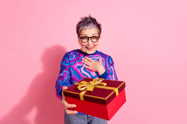照片肖像漂亮的退休女性兴奋地收到礼盒穿蓝色的彩色服装孤立在粉红色的背景图片下载