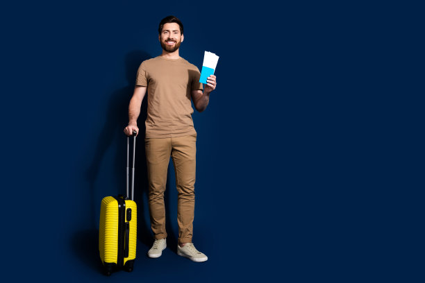 年轻帅哥的照片肖像，深色头发的模特，穿着米色t恤，拿着行李和机票去机场，在深蓝色的背景上孤立起来图片下载
