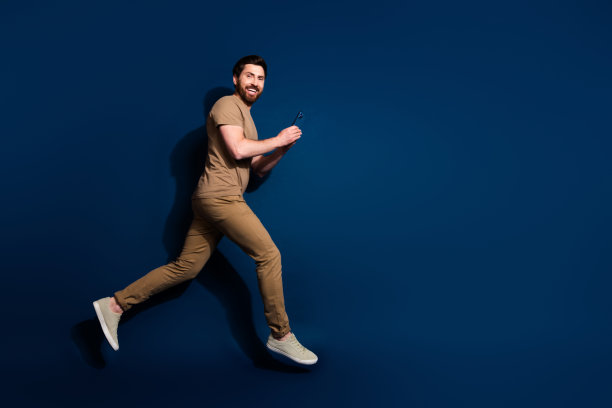照片年轻的跳跃家伙黑发模特在米色t恤使用跳跃与手机运行孤立的深蓝色背景图片下载