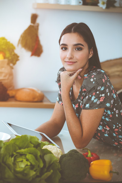 在阳光明媚的厨房里，一位年轻迷人的黑发女子坐在餐桌旁，正在挑选一顿美味佳肴的食谱图片下载