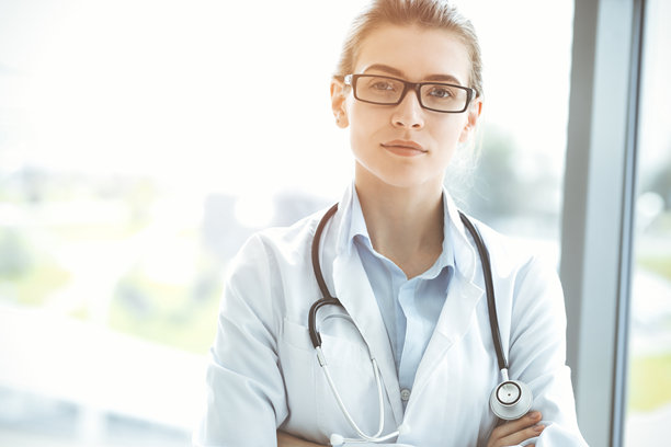 在阳光明媚的医院里，带着听诊器的美女医生微笑着，随时准备帮助病人。医学的概念图片下载