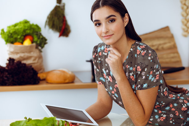 一位年轻迷人的黑发女子坐在厨房的餐桌旁，正在挑选一顿美味佳肴的食谱。平板电脑是最好的食谱图片下载