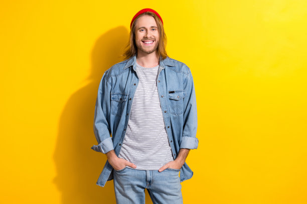 照片中，友好的帅哥留着长发，穿着牛仔裤衬衫，站在黄色背景上，双手放在口袋里图片下载