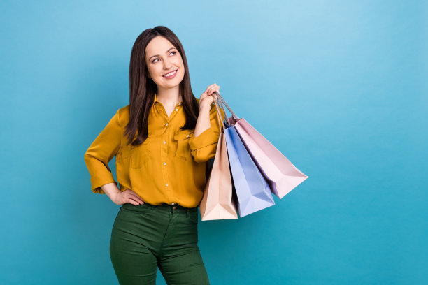 照片中迷人梦幻的女人穿着黄色衬衫抱着购物者看着空空荡荡的蓝色背景图片下载