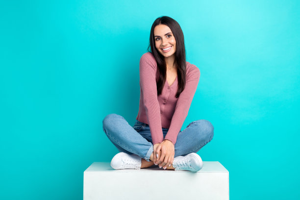 迷人的微笑女孩的全身照片，穿着时髦的衣服，盘腿放松地坐在白色的平台上，孤立在海蓝宝石的背景上图片下载