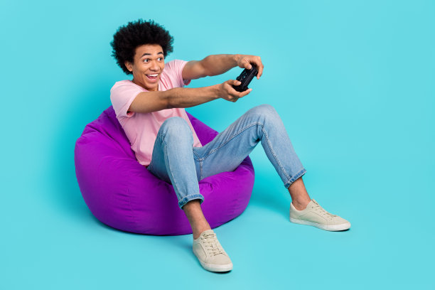 年轻的专业游戏机玩家的全身照片有趣的chevelure坐在豆袋游戏区广告持有操纵杆孤立的蓝色背景图片下载
