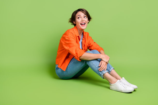 漂亮兴奋的女士穿着牛仔裤衬衫坐在地板上看空空间孤立的绿色背景的全长照片图片下载