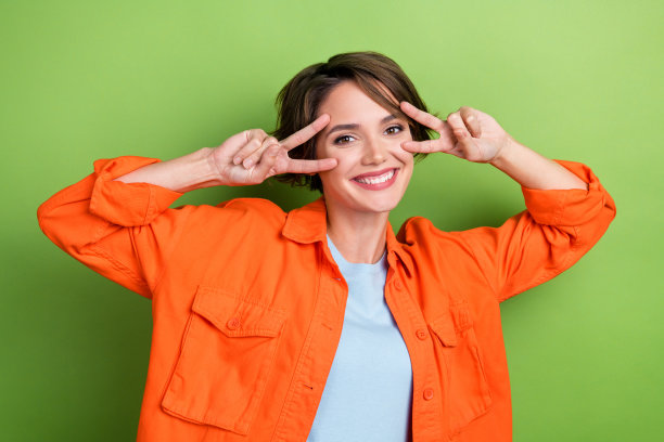 照片中，快乐可爱的漂亮女士穿着时髦的橙色衣服，两只手臂靠近脸，在绿色的背景下，v字标志好心情图片下载