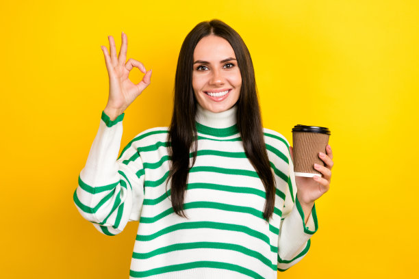 照片中可爱可爱的女人穿着条纹毛衣喝茶，显示出好的标志，孤立的黄色背景图片下载