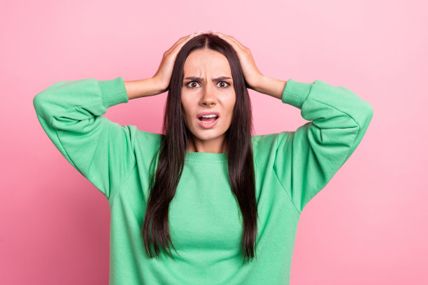 照片中，一头直发、身穿绿色运动衫的惊恐女子双手抱在头上，孤立在粉红色的背景上图片下载