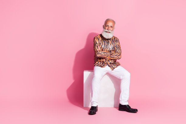 照片上的奢侈迷人的男人双臂交叉，穿着时髦的印花衣服，坐在空旷的地方，孤立在粉红色的背景上图片下载