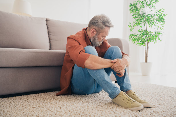 高级男性的全身照片坐在地板上沮丧地悲伤地抱着膝盖穿着棕色的衣服舒适的日光家室内客厅图片下载