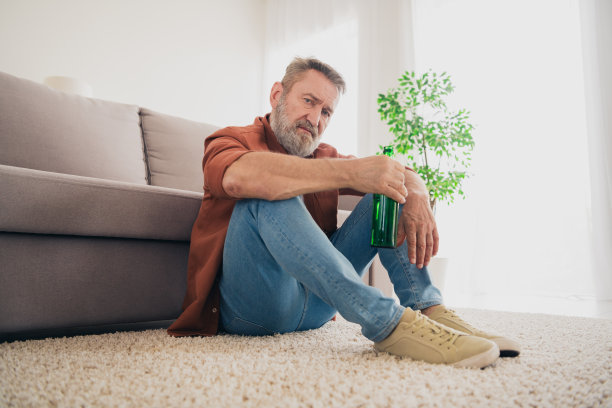 漂亮的退休老人男子的全尺寸照片，坐在地板上郁郁寡欢喝酒穿着棕色的衣服宽敞的房子室内的房间图片下载