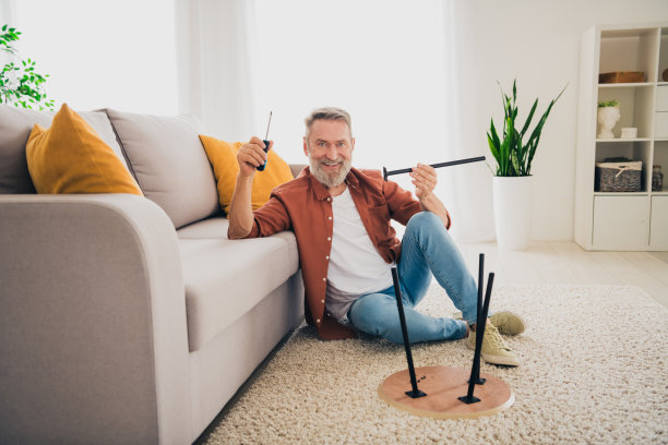 漂亮退休男子的全身照片坐在地板修理坏了的椅子腿螺丝刀穿棕色的衣服现代舒适的室内平坦的室内图片下载