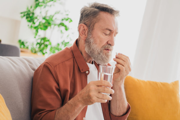 老年男性的照片坐在沙发上吃药喝水玻璃杯穿着棕色的衣服舒适的日光家室内客厅图片下载