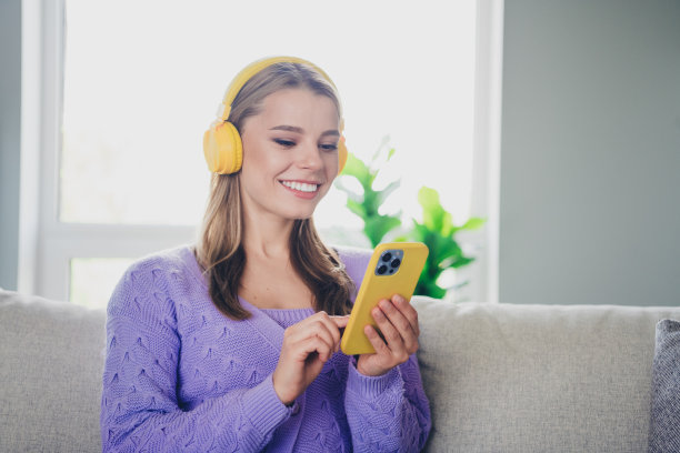 漂亮的年轻女孩的照片，坐在沙发上，耳机听音乐，拿着设备，穿着紫罗兰色的衣服，现代舒适的室内平坦的室内图片下载