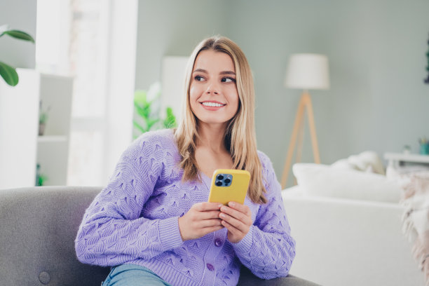 迷人的年轻女子的照片，坐在扶手椅，手持设备，看空的空间，穿着紫色的衣服舒适的日光家室内客厅图片下载