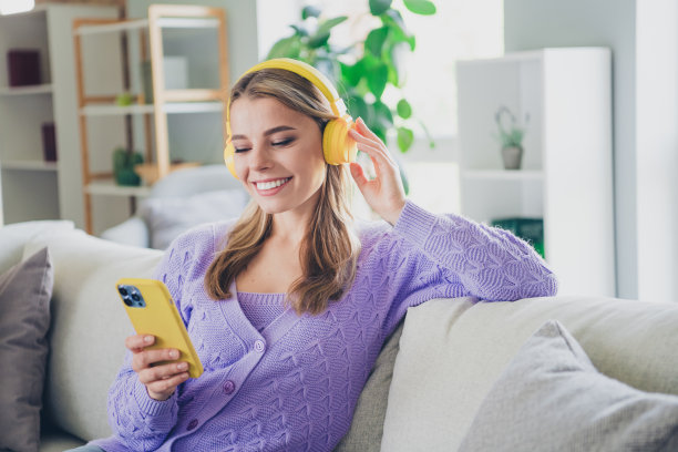 迷人的年轻女子的照片，坐在沙发上，耳机，手持设备，听音乐，穿着紫色的衣服，白天，家里室内客厅图片下载
