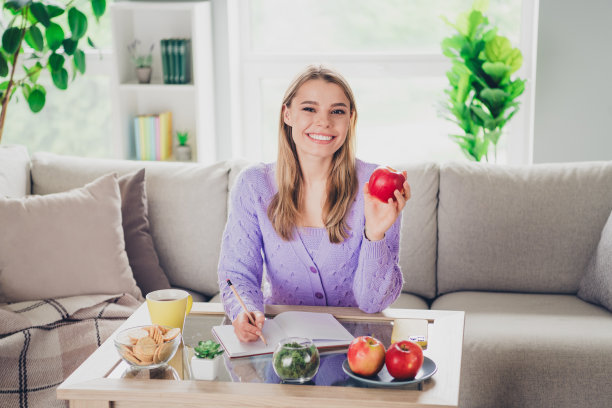 可爱开朗的女孩照片坐在沙发桌子写笔记本吃苹果穿着紫色的衣服度过消遣宽敞的房子在室内房间图片下载