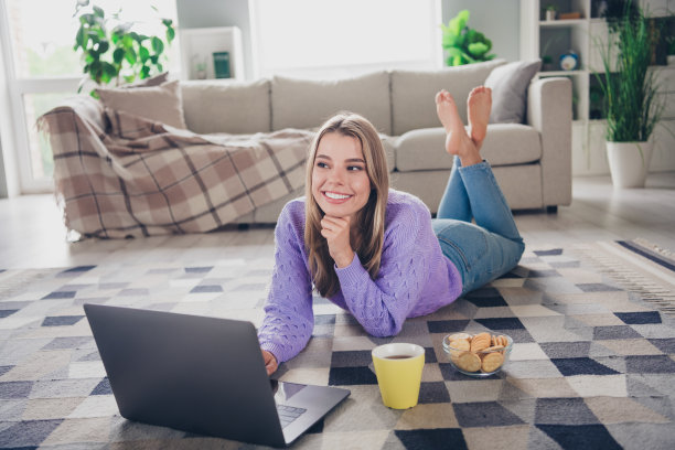 年轻女子躺在地板上的全尺寸照片上网本看电影零食放松穿着紫色的衣服舒适的日光家室内客厅图片下载