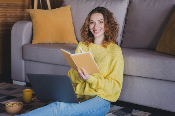 漂亮的年轻女子的照片，写笔记本，坐在地板上网本，穿着时尚的黄色毛衣，现代的光舒适的室内，室内平坦图片下载