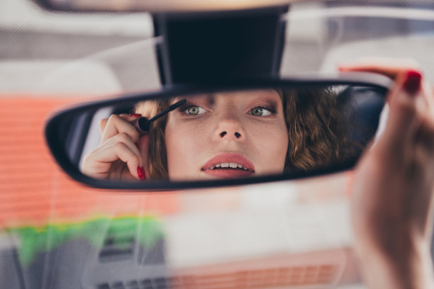 照片中，光鲜迷人的女司机穿着白衬衫，微笑着骑着车，在城市街道上涂睫毛膏图片下载