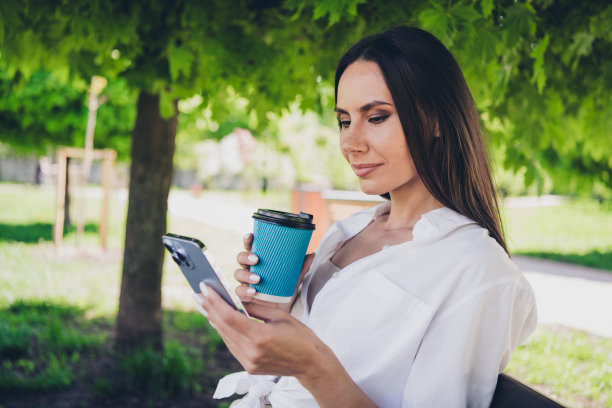 迷人的年轻开朗的女孩的照片，坐在长凳上喝咖啡，纸杯，拿着装置，穿着白色的衣服在外面玩图片下载