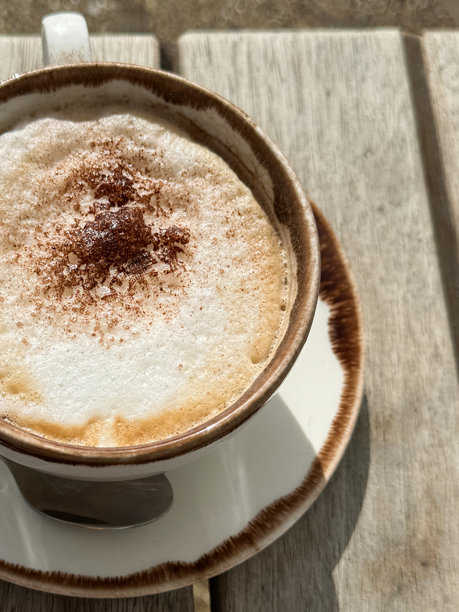 特写图片:刚倒入的卡布奇诺咖啡，装在白色的杯子和茶托上，茶托上放着茶勺，牛奶上撒着巧克力粉，热饮，阳光明媚的户外用餐环境，高处的景色图片下载