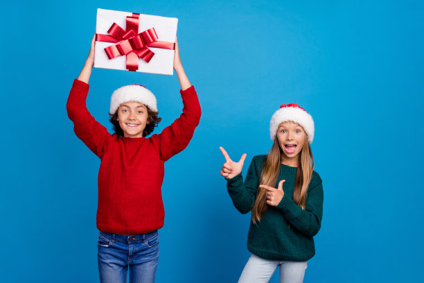 照片中的两人孩子，男孩举起礼盒，女孩点圣诞孤立的蓝色背景图片下载
