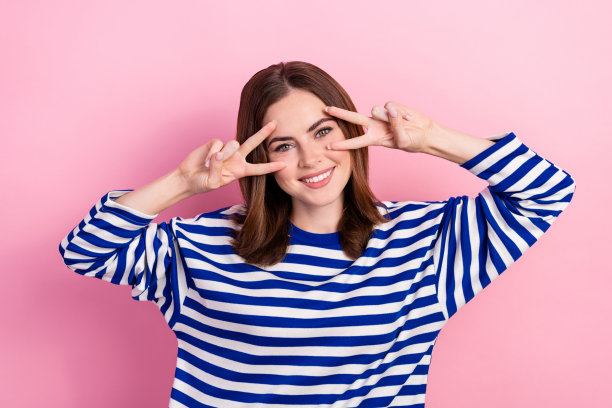 快乐漂亮的女士的特写照片，穿着五颜六色的衣服，手臂近脸，在粉红色的背景上展示v字图片下载