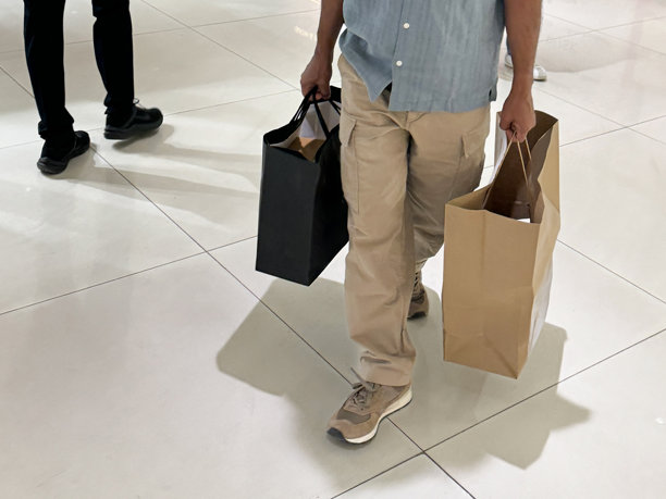 一个无法辨认的男人走过零售商店的服装部，提着装满商品的纸袋，重点是前景、消费主义和购物理念图片下载