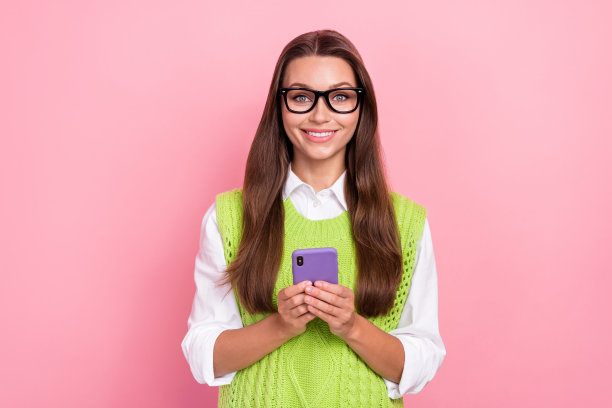 照片中，可爱的女士穿着时髦的衣服，拿着iphone三星，在粉红色的背景上宣传现代设备图片下载