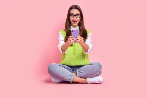 开朗积极的女孩的全身照片直发绿色背心看智能手机阅读信息孤立在粉红色的背景图片下载