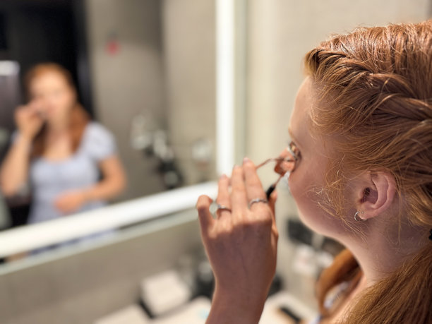 特写照片:迷人的红发女子，梳着辫子，穿着背心裙，站在浴室的水池边，镜子里的倒影，用睫毛夹，涂化妆品，侧影，自我护理和美容概念图片下载