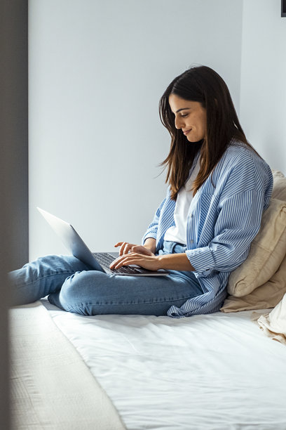 一位年轻的职业女性舒适地坐在酒店的床上，用一台带有无线wifi连接的笔记本电脑进行远程工作。捕捉了数字游牧民的现代生活方式，他们在网上出差和工作图片下载