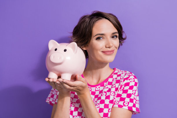 可爱的年轻女士的照片肖像拿着存钱的小猪穿着时尚的粉红色服装孤立的紫色背景图片下载