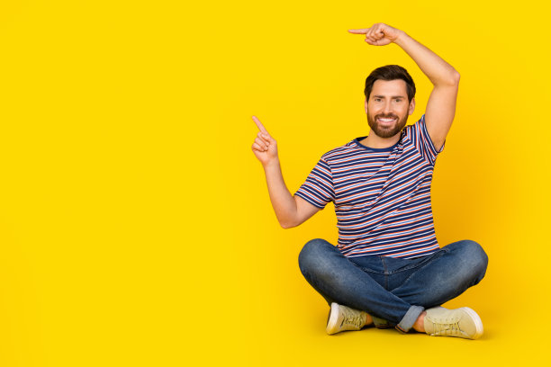 穿着休闲服的年轻人全身照片，坐在地板上，手指指向空白的空间，在黄色背景上孤立的瑜伽学校广告图片下载