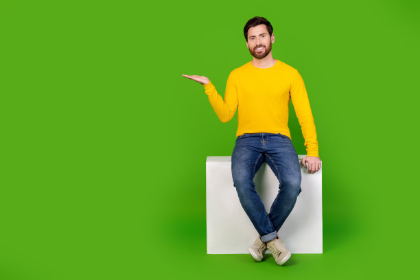 漂亮的年轻男性的全尺寸照片，抱着空旷的空间坐着立方体，在绿色的背景上穿时髦的黄色衣服图片下载