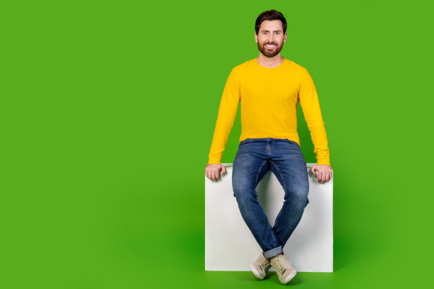 全身照片的迷人的年轻人坐在白色立方体空的空间穿着时尚的黄色衣服孤立在绿色的背景图片下载