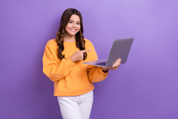 照片中可爱的女孩穿着橙色运动衫，指着苹果三星现代设备孤立的紫色背景图片下载