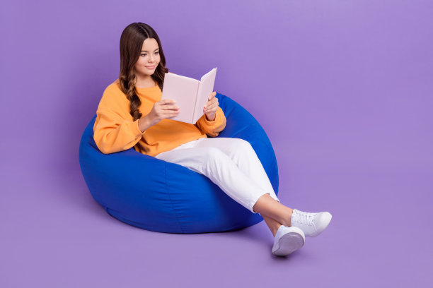 甜美漂亮的女士穿着橙色运动衫豆袋享受书空空间孤立的紫罗兰色背景的全身照片图片下载